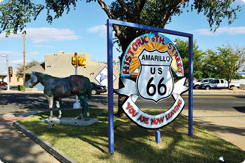 Ein Schild mit der Aufschrift „Historic 6th Street Amarillo US 66 You Are Now Leaving“ steht neben einer Stra enkreuzung, auf der eine Pferdeskulptur auf einer kleinen Rasenfl che steht. Beschreibung von KI generiert