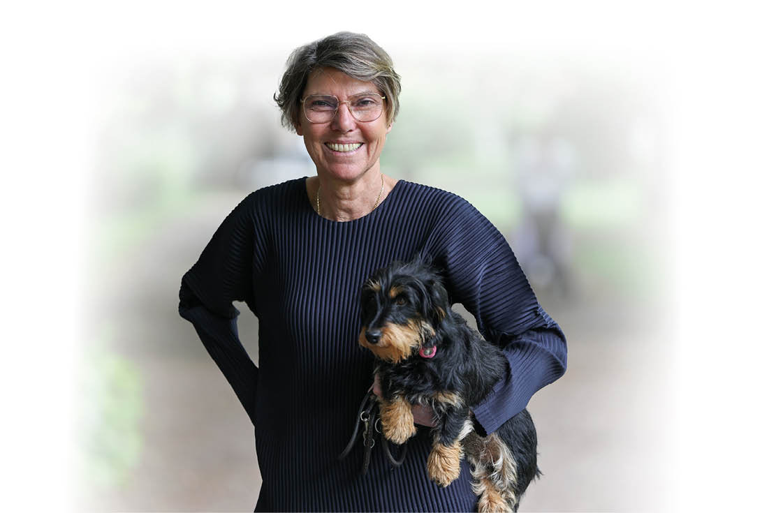 Eine l chelnde Person mit kurzen grauen Haaren und Brille tr gt einen kleinen schwarzen Hund mit braunen Abzeichen und steht vor einem unscharfen Hintergrund. Beschreibung von KI generiert