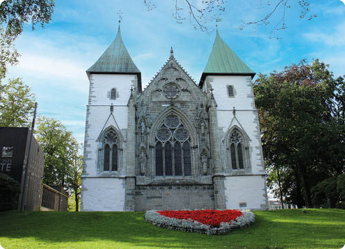 Eine historische Kirche mit zwei spitzen T rmen und einem gro en, verzierten Fenster in der Mitte steht auf einem gr nen H gel mit einem Blumenbeet in Herzform davor. Beschreibung von KI generiert