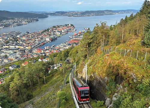 Eine rote Standseilbahn f hrt auf einer schmalen Strecke durch bewaldetes, h geliges Gel nde mit Blick auf eine Stadt und einen gro en Fjord im Hintergrund. Beschreibung von KI generiert