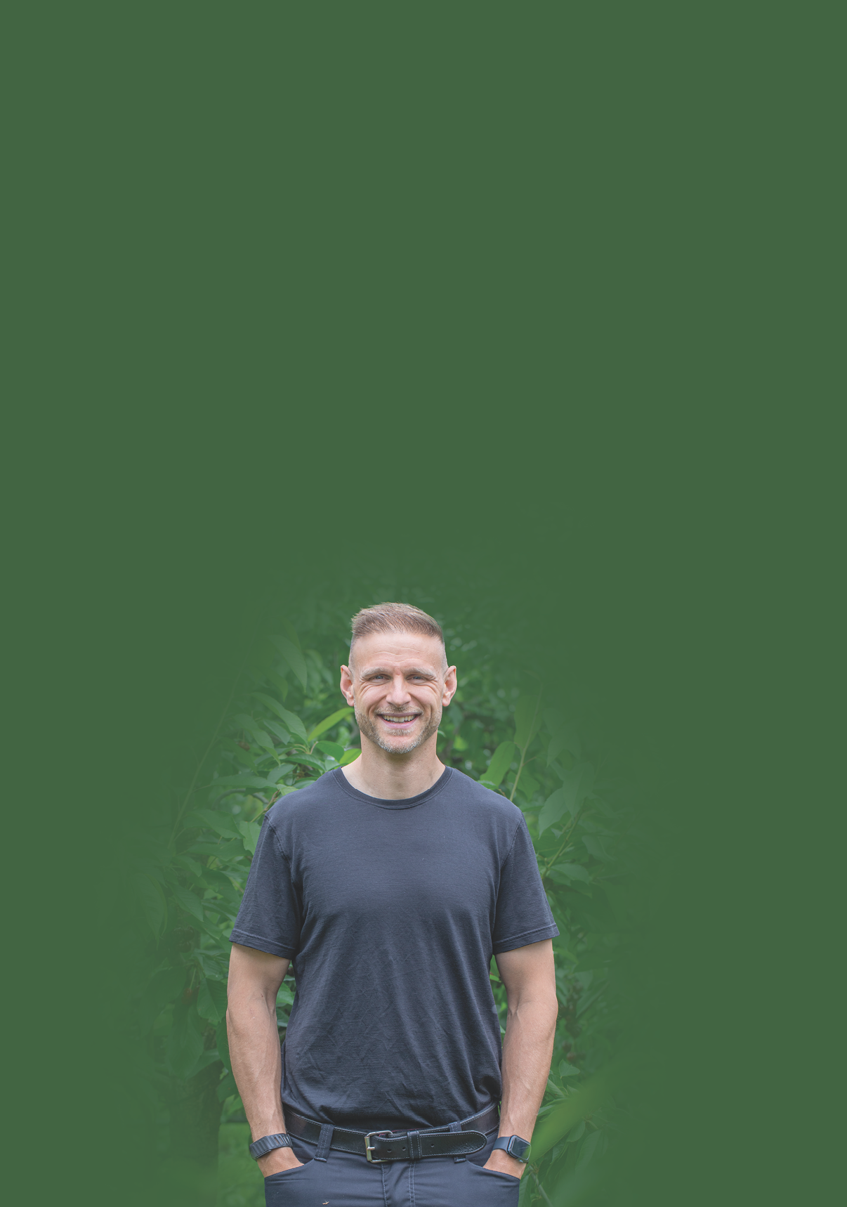 Eine l chelnde Person mit kurzen Haaren tr gt ein dunkles T-Shirt und eine Armbanduhr und steht vor einem gr nen, unscharfen Hintergrund mit Pflanzen. Beschreibung von KI generiert