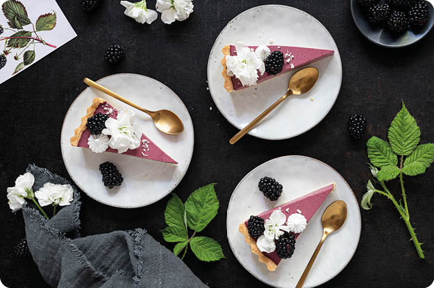 Drei Teller mit je einem St ck Brombeertorte, garniert mit wei en Bl ten und frischen Brombeeren, liegen auf einem dunklen Untergrund neben gr nen Bl ttern, einem grauen Stoff und einer kleinen Schale mit Brombeeren. Beschreibung von KI generiert