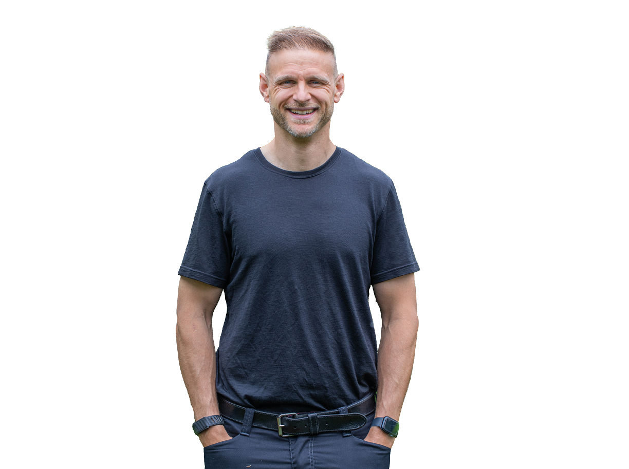 Eine l chelnde Person mit kurzen Haaren tr gt ein dunkles T-Shirt und hat die H nde in den Hosentaschen. Beschreibung von KI generiert