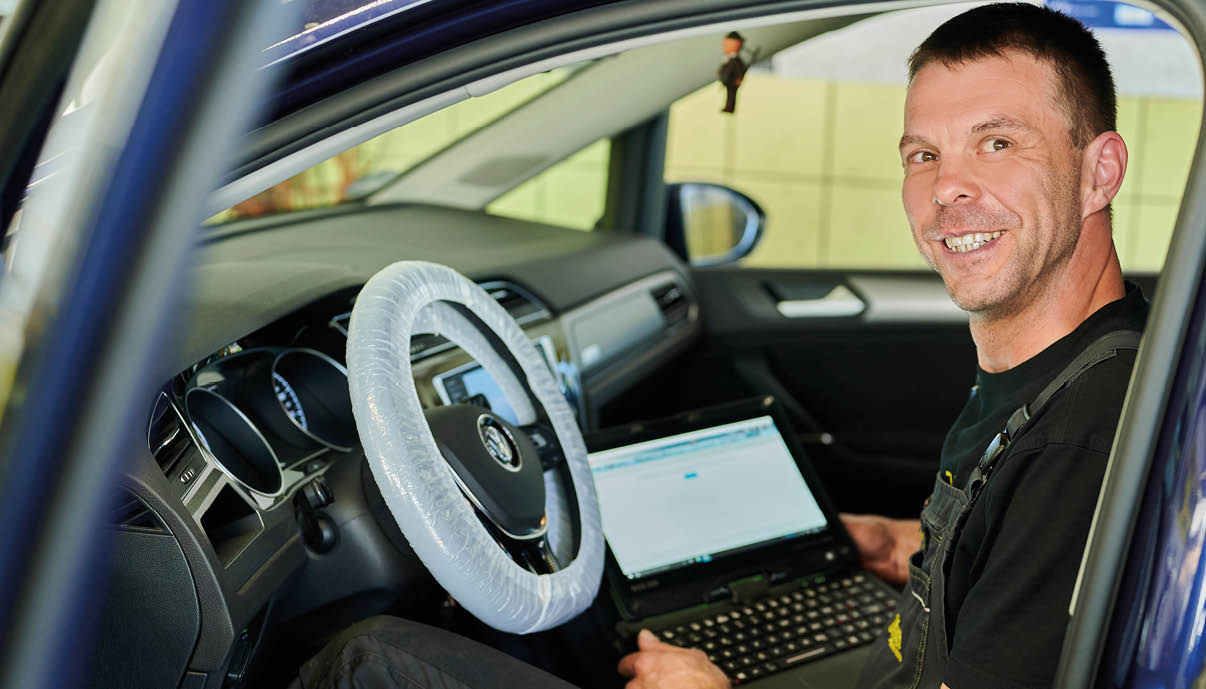 Eine Person in Arbeitskleidung sitzt l chelnd im Fahrersitz eines Autos und arbeitet an einem Laptop, der auf dem Scho liegt.  Beschreibung von KI generiert