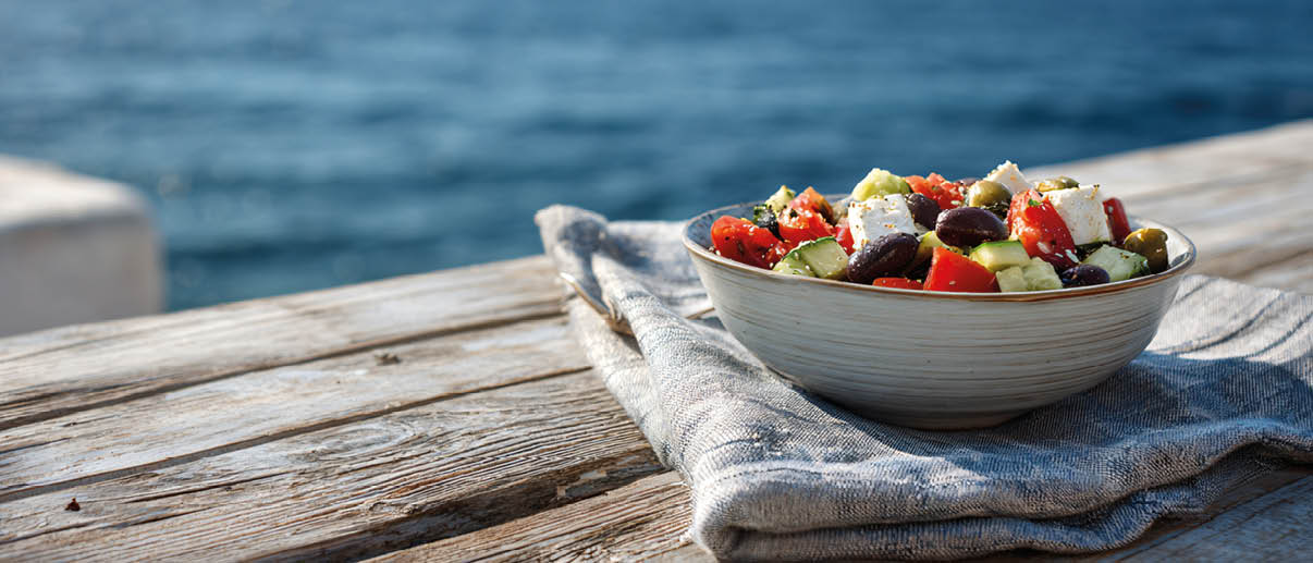 Eine Sch ssel mit einem bunten Salat aus Tomaten, Gurken, Oliven und Feta steht auf einem grauen Tuch auf einem h lzernen Tisch mit Blick auf das Meer im Hintergrund. Beschreibung von KI generiert