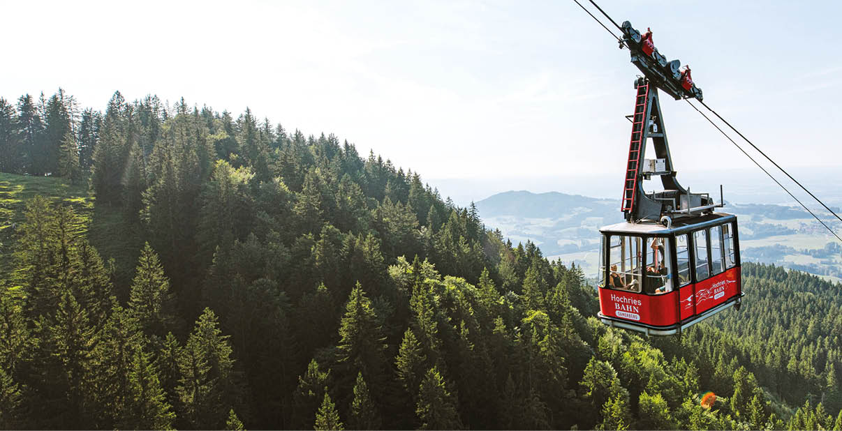 Eine rote Seilbahn f hrt ber einen bewaldeten Berghang mit weiter Landschaft im Hintergrund.  Beschreibung von KI generiert