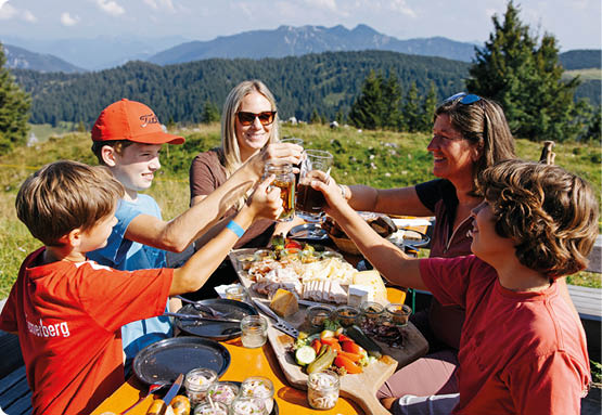 F nf Personen sitzen an einem Holztisch im Freien in einer bergigen Landschaft und sto en mit Getr nken an, w hrend sie gemeinsam essen und lachen. Beschreibung von KI generiert
