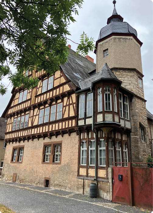 Ein historisches Fachwerkhaus mit einem runden Turm und vielen Fenstern steht an einer gepflasterten Stra e neben einem Baum. Beschreibung von KI generiert