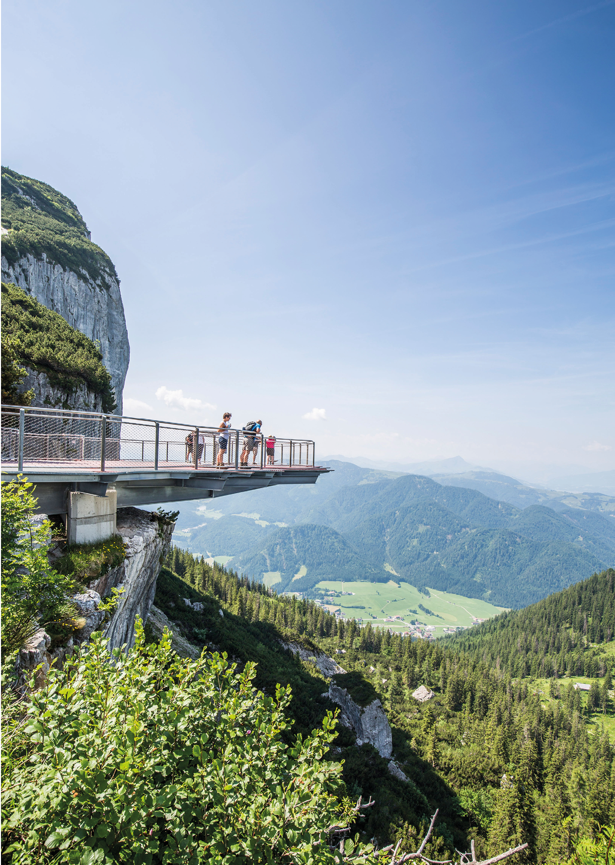 Mehrere Personen stehen auf einer Aussichtsplattform, die an einer steilen Felswand ber einem bewaldeten Tal mit Bergen im Hintergrund befestigt ist.  Beschreibung von KI generiert
