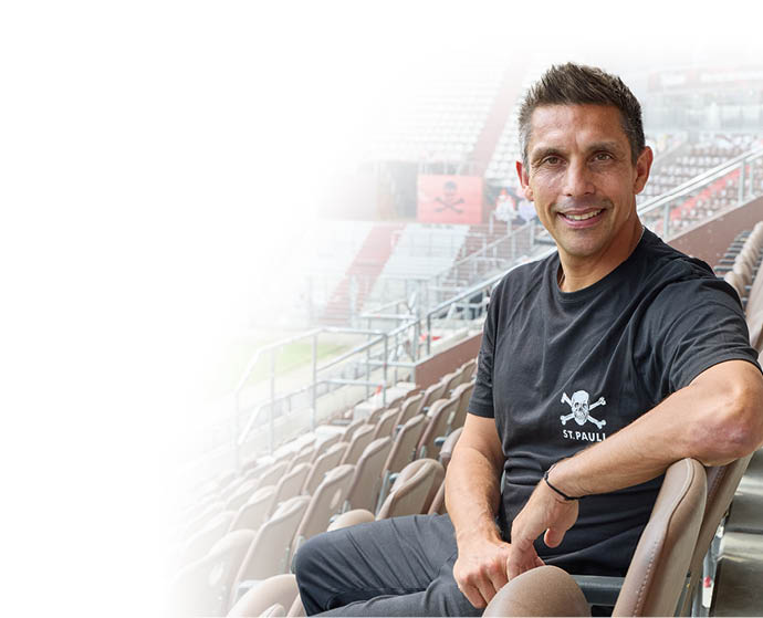 Eine Person mit kurzem Haar tr gt ein schwarzes T-Shirt mit Totenkopf-Logo und sitzt l chelnd auf einem Sitzplatz in einem fast leeren Stadion. Beschreibung von KI generiert