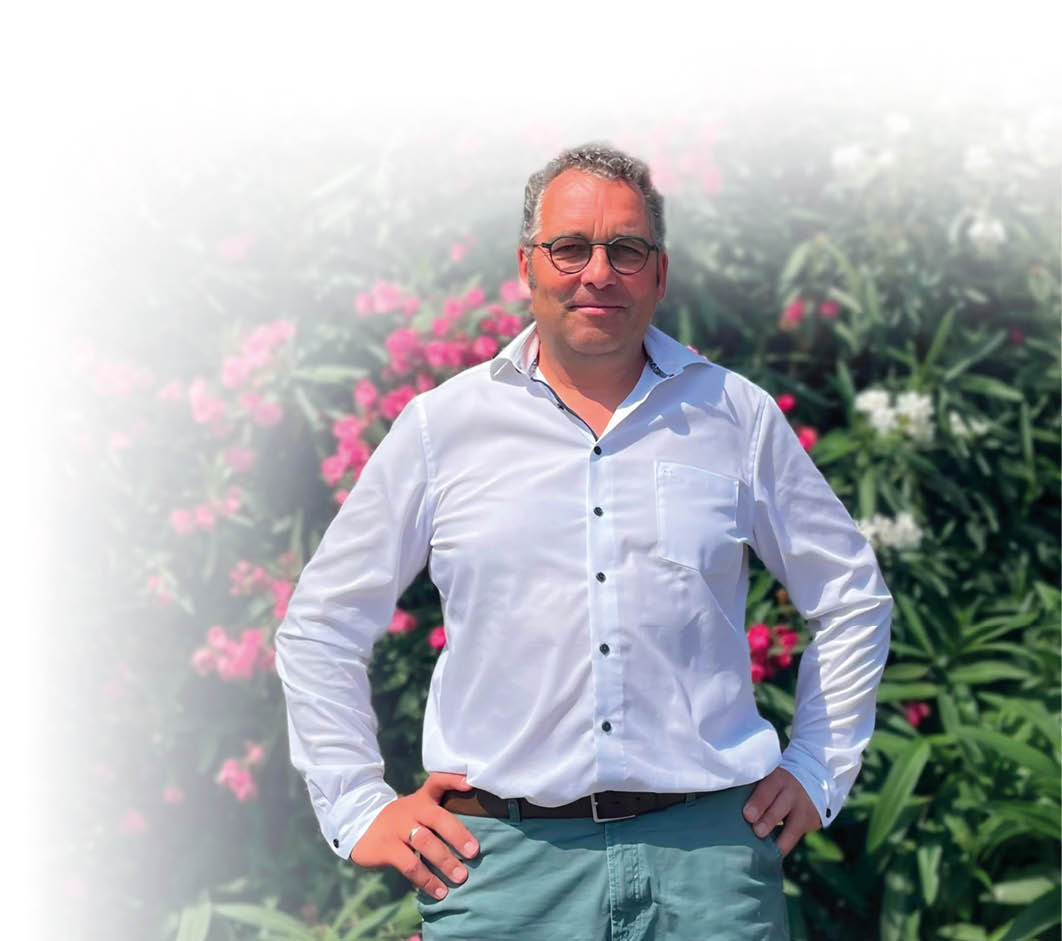 Eine Person mit Brille tr gt ein wei es Hemd und steht mit den H nden in den H ften vor einem Hintergrund aus gr nen Pflanzen und rosa Bl ten. Beschreibung von KI generiert