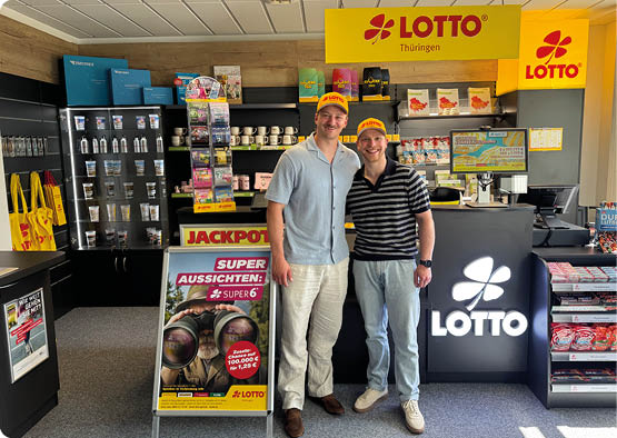 Zwei Erwachsene mit gelben Lotto-M tzen stehen l chelnd in einem Lotto-Laden vor einem beleuchteten Lotto-Schalter und Werbeaufstellern. Beschreibung von KI generiert