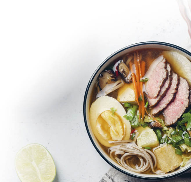 Eine Sch ssel mit einer asiatisch inspirierten Suppe mit Nudeln, Gem se, Kr utern, Ei und Fleisch, daneben liegen Essst bchen und eine Stoffserviette. Beschreibung von KI generiert