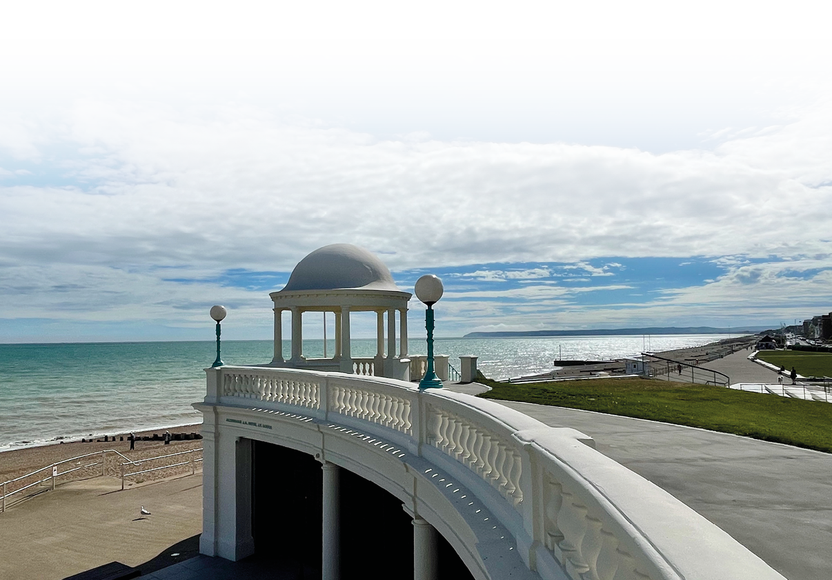 Eine wei e, elegante Strandpromenade mit einer kleinen Kuppelterrasse erstreckt sich entlang eines ruhigen Meeres unter einem teilweise bew lkten Himmel. Beschreibung von KI generiert
