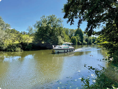 Ein Boot f hrt auf einem ruhigen Fluss, umgeben von gr nen B umen und unter einem klaren blauen Himmel. Beschreibung von KI generiert