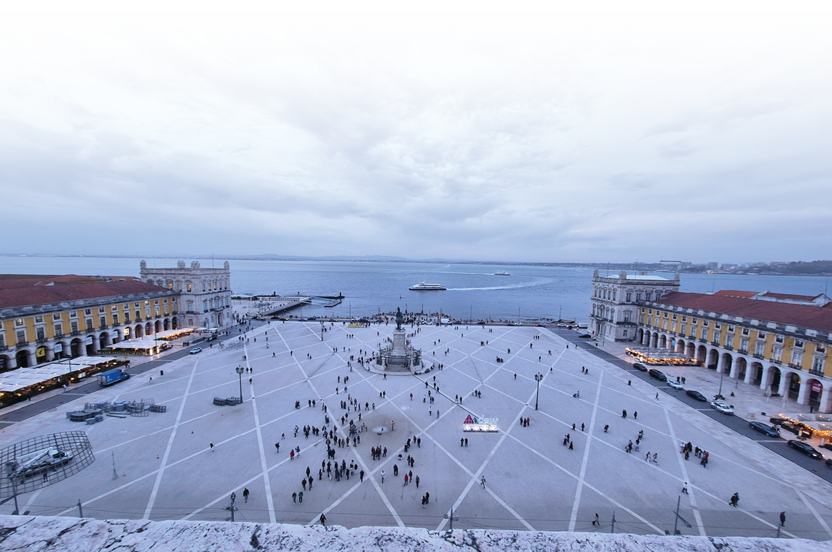 Ein gro er offener Platz mit einem zentralen Denkmal ist von historischen Geb uden mit Arkaden umgeben und im Hintergrund ist das Meer mit mehreren Booten unter einem bew lkten Himmel zu sehen. Beschreibung von KI generiert
