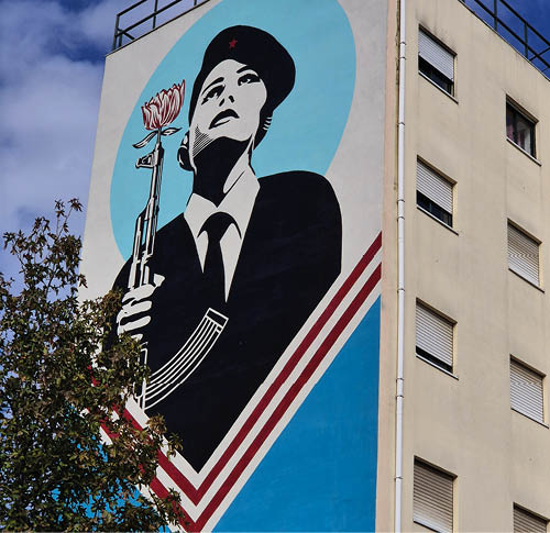 An der Seite eines Geb udes ist ein gro es Wandbild in einem grafischen Stil zu sehen, das eine Person in Uniform mit einer M tze zeigt, die eine Blume h lt, vor einem hellblauen Hintergrund mit roten und wei en Streifen. Beschreibung von KI generiert