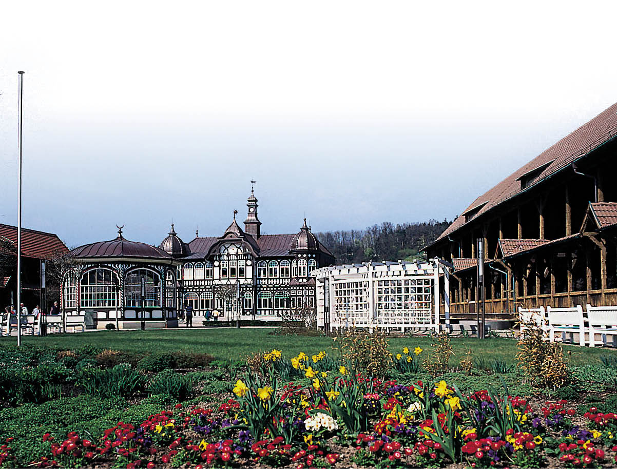 Ein historisches Geb ude mit aufwendiger Holzarchitektur steht hinter einem bunten Blumenbeet und einer gr nen Wiese unter einem bew lkten Himmel. Beschreibung von KI generiert