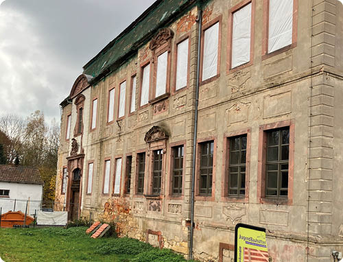 Ein altes, teilweise verfallenes Geb ude mit zugemauerten Fenstern steht neben einer Wiese, auf der ein Werbebanner mit einer Person in traditioneller Kleidung steht. Beschreibung von KI generiert