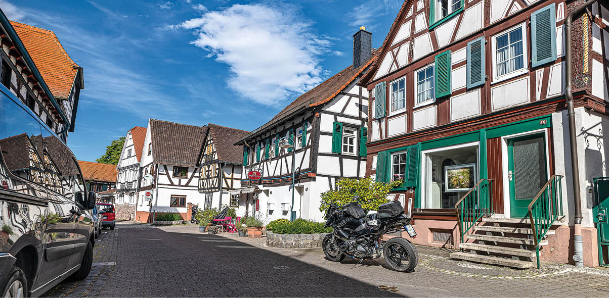 Eine ruhige Stra e mit Fachwerkh usern, einem Motorrad und einem geparkten Auto unter einem blauen Himmel mit einigen Wolken. Beschreibung von KI generiert