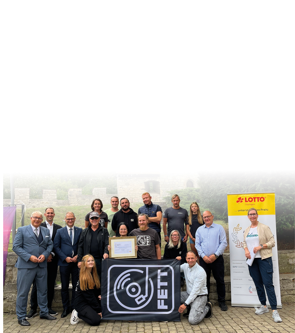 Eine Gruppe von Erwachsenen posiert l chelnd im Freien vor einer Steinmauer mit einem gro en Banner und einem aufgestellten Werbeplakat, wobei eine Person eine Urkunde h lt. Beschreibung von KI generiert