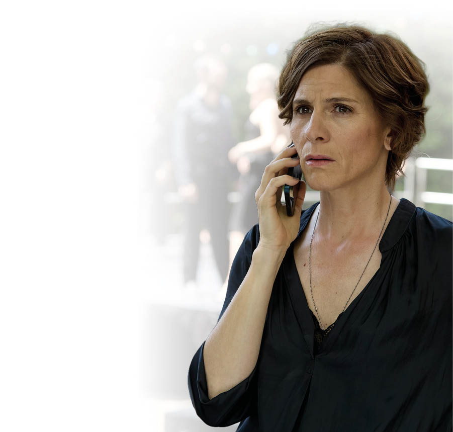 Eine Person mit kurzen braunen Haaren tr gt eine schwarze Bluse und spricht besorgt in ein Mobiltelefon, w hrend im Hintergrund zwei weitere Personen unscharf zu sehen sind. Beschreibung von KI generiert