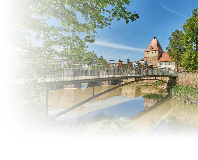 Mehrere Personen mit Fahrr dern stehen auf einer Br cke ber einem ruhigen Fluss vor einem historischen Geb ude mit rotem Dach und blauem Himmel.  Beschreibung von KI generiert