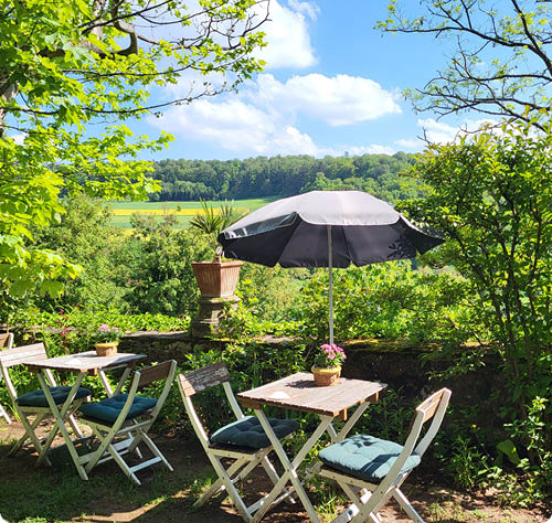 Ein sonniger Garten mit zwei kleinen Tischen, St hlen mit Kissen und einem gro en Sonnenschirm vor einer gr nen Landschaft und blauem Himmel. Beschreibung von KI generiert