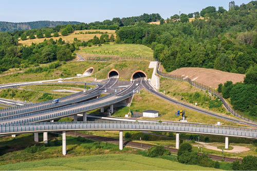 Mehrspurige Autobahnen f hren in mehrere Tunnel, die in einer gr nen, h geligen Landschaft eingebettet sind. Beschreibung von KI generiert