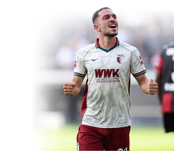 Ein jubelnder Spieler in wei -rotem Fu balltrikot steht auf dem Spielfeld, w hrend eine weitere Person in schwarzem Trikot mit der Nummer drei im Hintergrund zu sehen ist. Beschreibung von KI generiert