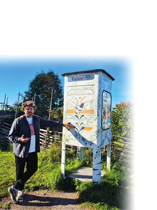 Eine Person mit Kranz auf dem Kopf steht neben einer bemalten Telefonzelle mit floralen Motiven und zeigt darauf, w hrend sie auf einem Weg in einer gr nen, l ndlichen Umgebung steht. Beschreibung von KI generiert
