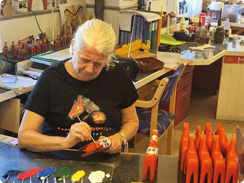Eine Seniorin mit hellen Haaren sitzt an einem Tisch und bemalt sorgf ltig eine kleine rote Holzfigur, w hrend mehrere hnliche Figuren und Farbt pfe vor ihr liegen.  Beschreibung von KI generiert