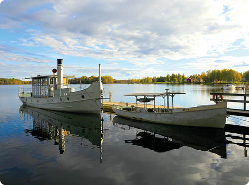Zwei Boote liegen an einem Steg auf einem ruhigen See mit herbstlich gef rbten B umen im Hintergrund unter einem teilweise bew lkten Himmel. Beschreibung von KI generiert