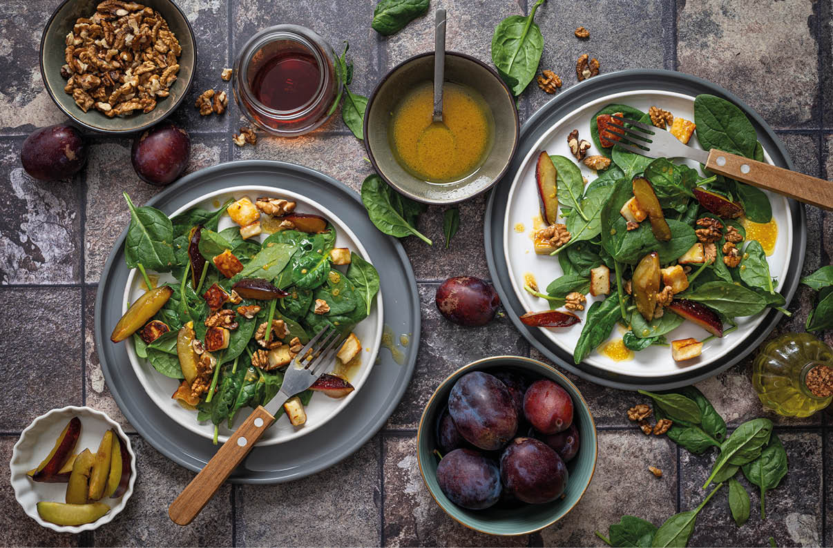 Zwei Teller mit frischem Spinatsalat, Waln ssen, gebratenem K se und getrockneten Chilischoten sind auf einem Steinfliesenuntergrund zusammen mit Schalen voller Pflaumen, Waln ssen, einem Glas Honig und einer Schale mit Dressing angerichtet. Beschreibung von KI generiert