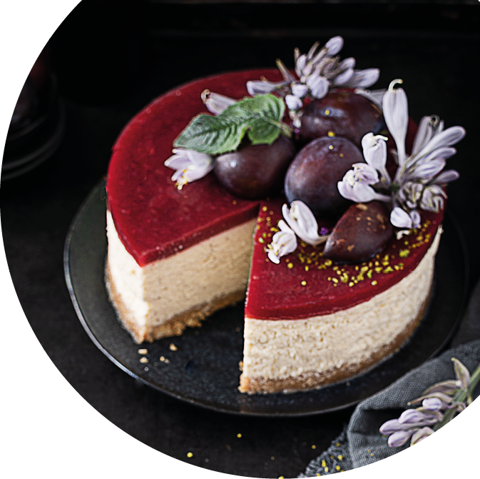 Ein cremiger Kuchen mit roter Fruchtschicht oben ist auf einem schwarzen Teller angerichtet und mit frischen Feigen, gr nen Bl ttern und wei en Bl ten dekoriert. Beschreibung von KI generiert
