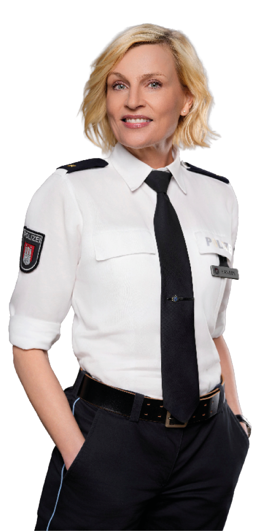 Eine Person mit kurzen blonden Haaren tr gt eine wei e Uniformbluse mit schwarzen Schulterklappen und Krawatte und steht mit den H nden in den Hosentaschen vor wei em Hintergrund. Beschreibung von KI generiert