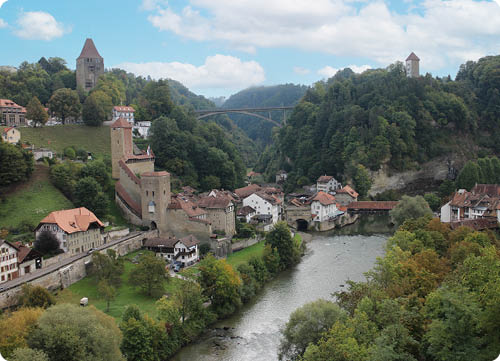 Eine historische Stadt mit alten T rmen, Fachwerkh usern und einer Br cke ber einen Fluss, umgeben von bewaldeten H geln unter einem bew lkten Himmel.  Beschreibung von KI generiert