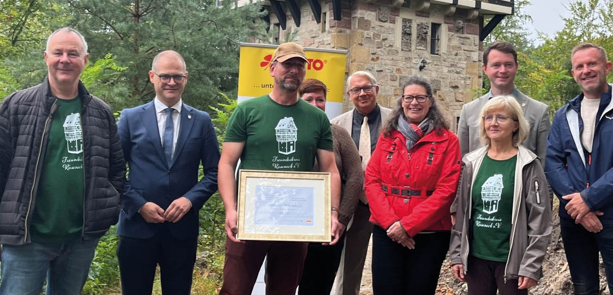 Eine Gruppe von acht Personen steht vor einem historischen Fachwerkhaus, wobei eine Person in der Mitte eine gerahmte Urkunde h lt und mehrere Personen gr ne T-Shirts mit einem wei en Logo tragen. Beschreibung von KI generiert
