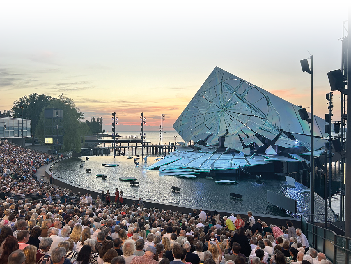Vor einer gro en Menschenmenge sitzt ein Publikum in einem Freilufttheater vor einer B hne mit einem modernen, spiegelnden B hnenbild, das wie zerbrochenes Glas aussieht, w hrend die Sonne am Horizont untergeht. Beschreibung von KI generiert