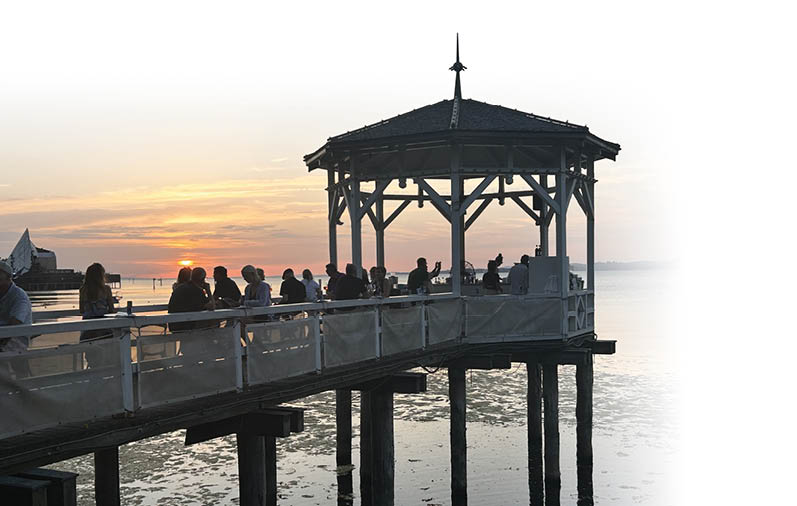 Mehrere Personen sitzen auf einem Steg mit einem berdachten Pavillon und beobachten den Sonnenuntergang  ber dem Wasser.  Beschreibung von KI generiert
