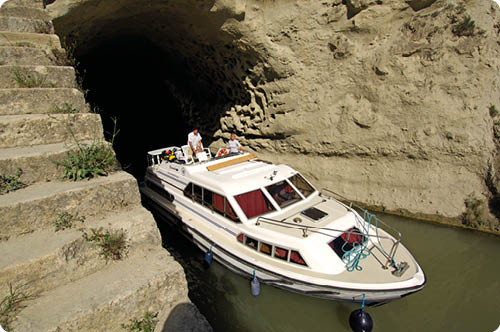 Ein Boot mit zwei Personen f hrt aus einem dunklen Tunnel in einem felsigen Gew sser heraus. Beschreibung von KI generiert
