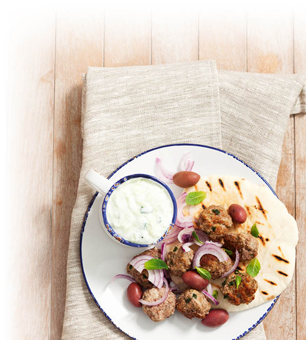Auf einem wei en Teller mit blauem Rand liegen gegrillte Fleischb llchen, rote Zwiebelringe, Oliven, Minzbl tter und ein Fladenbrot neben einer kleinen Schale mit wei em Dip auf einem hellen Leinentuch auf einem Holztisch. Beschreibung von KI generiert