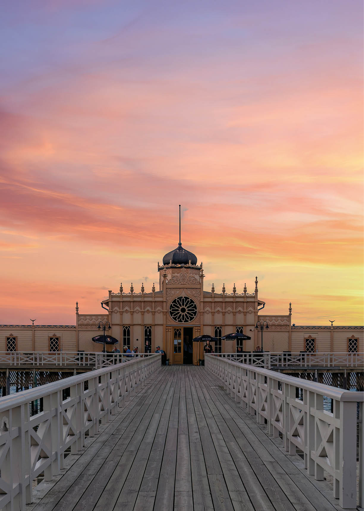 Ein historisches Geb ude auf Stelzen steht am Ende eines langen Holzstegs ber dem Wasser bei einem farbenfrohen Sonnenuntergang.  Beschreibung von KI generiert