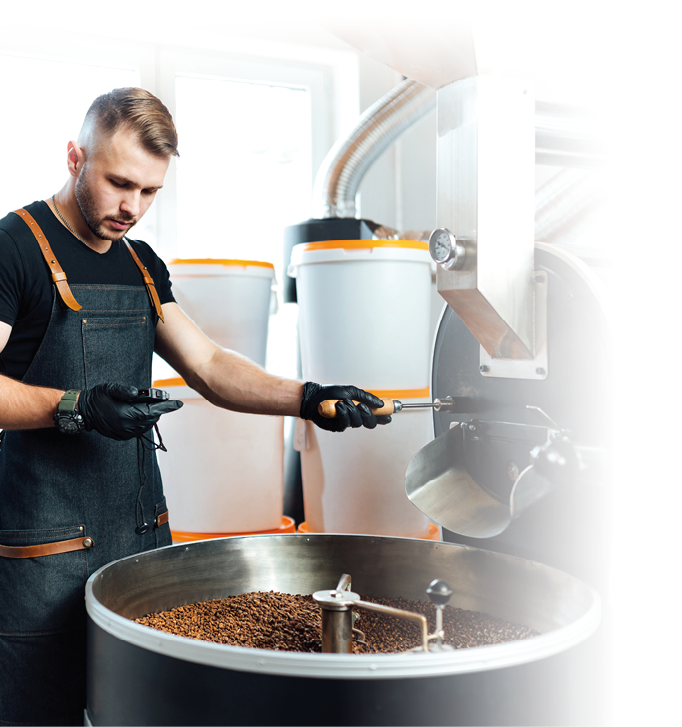 Eine Person mit Sch rze und Handschuhen berpr ft ger stete Kaffeebohnen in einer industriellen R stmaschine in einem hellen Raum.  Beschreibung von KI generiert