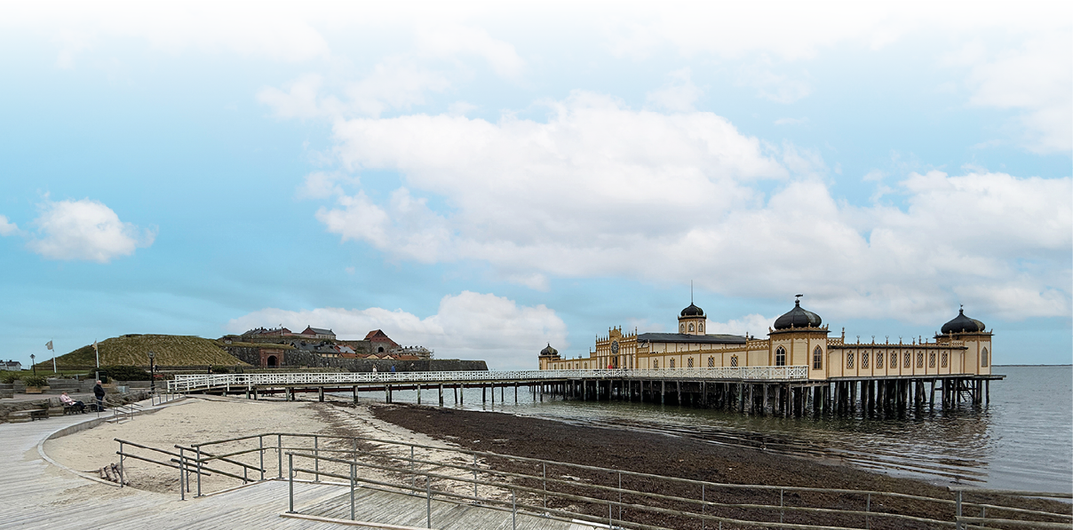 Ein historisches Geb ude auf Stelzen erstreckt sich ber das Meer an einem bew lkten Tag mit einem Sandstrand und einem befestigten Weg im Vordergrund.  Beschreibung von KI generiert