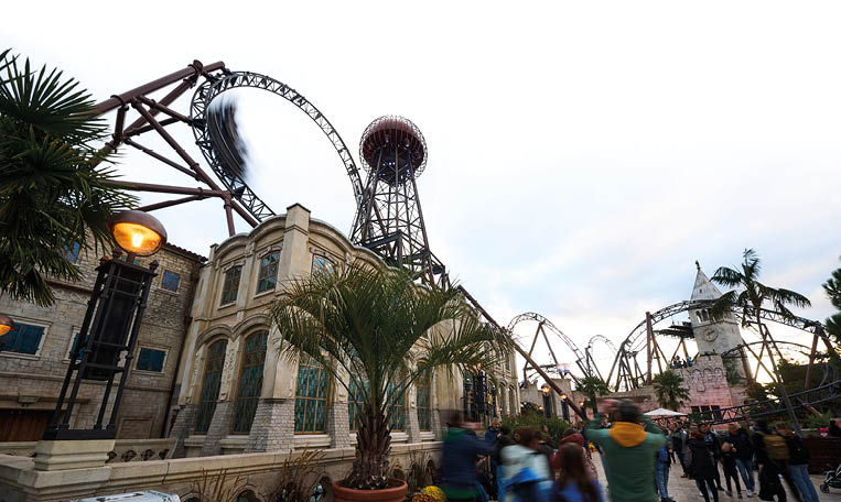 Ein belebter Freizeitpark mit einer Achterbahn, herbstlicher Dekoration aus K rbissen und Blumen sowie mehreren Menschen, die sich auf einem gepflasterten Weg bewegen. Beschreibung von KI generiert