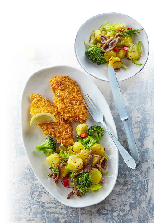 Zwei panierte Fischfilets mit Zitronenscheibe und ein bunter Gem sesalat mit Brokkoli, Kartoffeln und roten Paprikast cken sind auf zwei wei en Tellern angerichtet, daneben liegen Messer und Gabel. Beschreibung von KI generiert