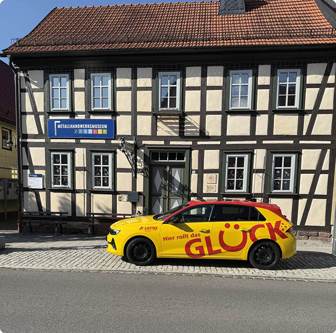 Ein gelbes Auto mit roter Aufschrift steht vor einem Fachwerkhaus mit einem Schild, das auf ein Museum hinweist. Beschreibung von KI generiert