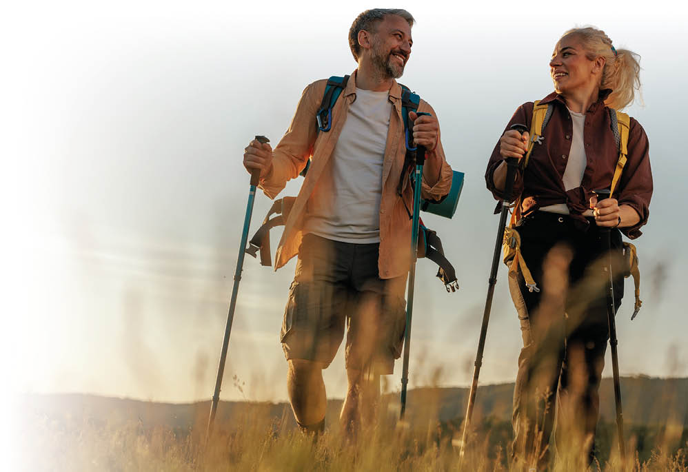 Zwei Erwachsene mit Wanderst cken und Rucks cken gehen l chelnd durch ein hohes Grasfeld bei Sonnenuntergang. Beschreibung von KI generiert