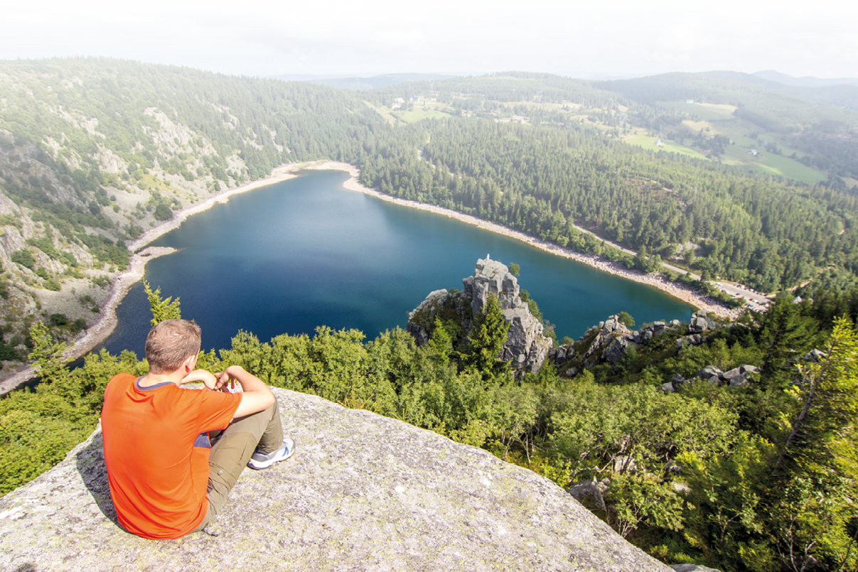 Eine Person in einem orangefarbenen T-Shirt sitzt auf einem Felsen und blickt auf einen von W ldern umgebenen See in einer h geligen Landschaft. Beschreibung von KI generiert